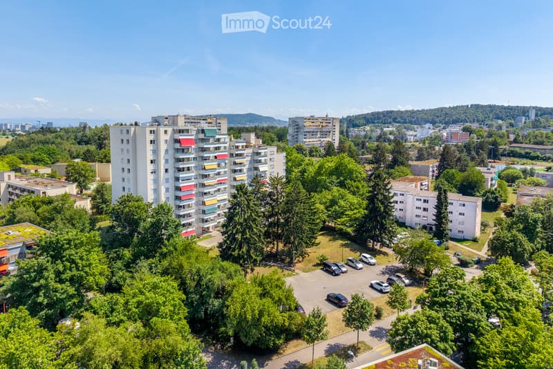 Aerial view of a residential area with multiple apartment buildings, surrounded by greenery and trees, with a parking lot and several parked cars.