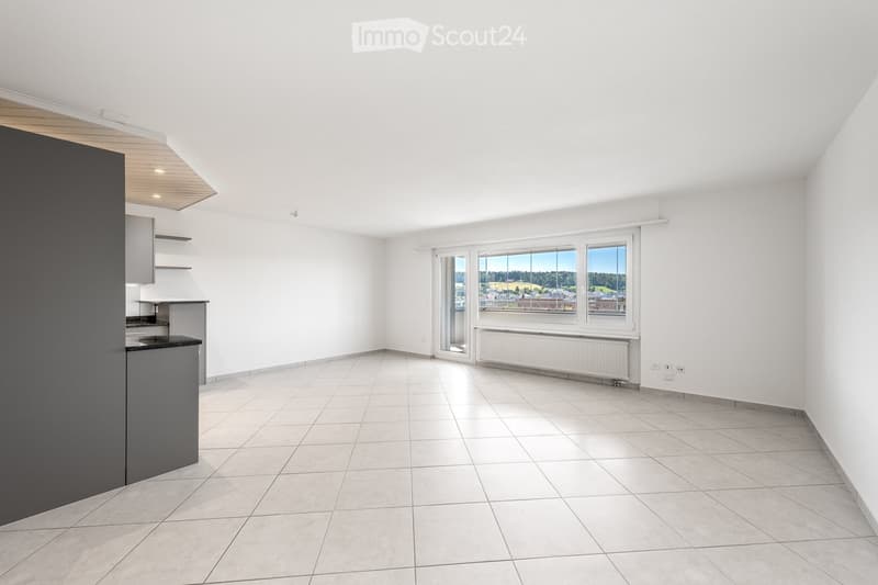 Empty room with white tiled flooring, white walls, and large windows offering a scenic view. The room features a small kitchen area with cabinets and a countertop on the left side.