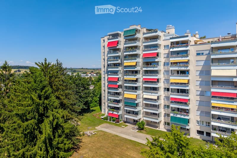 A modern multi-story apartment building with balconies, colorful awnings, and surrounding greenery.