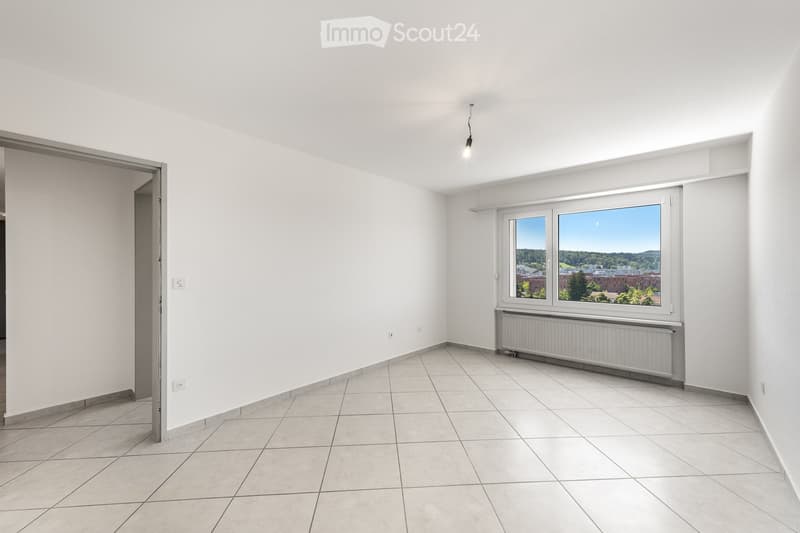 Empty room with white walls, tiled floor, large window with view, radiator, and single light fixture.