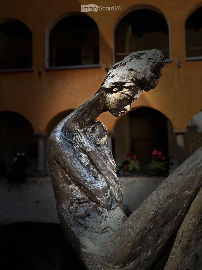 Bronze sculpture of a human figure in a dynamic pose, set against a backdrop of a historic building with arched windows and potted plants.