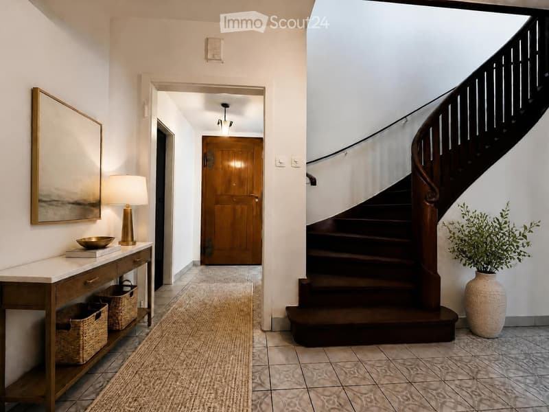 Elegant hallway with a wooden staircase, white walls, a console table with a lamp, and a large mirror. The floor is tiled, and there is a vase with greenery on the right side.