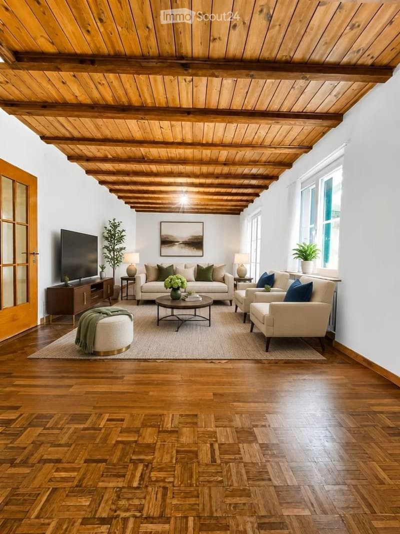 Modern living room with wooden floors, exposed wooden ceiling beams, large windows, and a mix of neutral and blue tones. Features a sofa, coffee table, ottoman, armchair, side tables, lamps, and a television.
