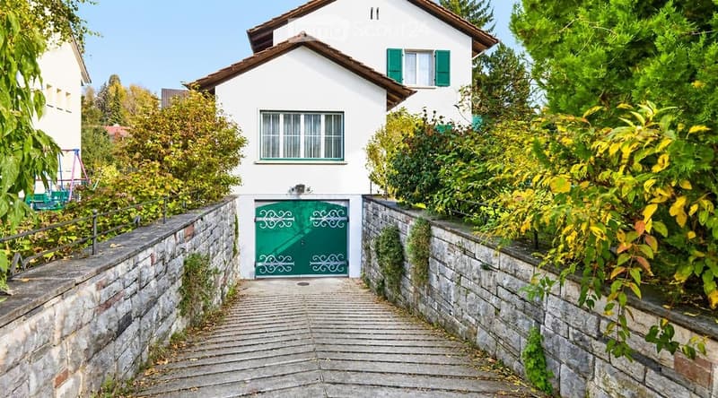 A two-story white house with green shutters and a green front door, surrounded by lush greenery and a stone pathway leading to the entrance.