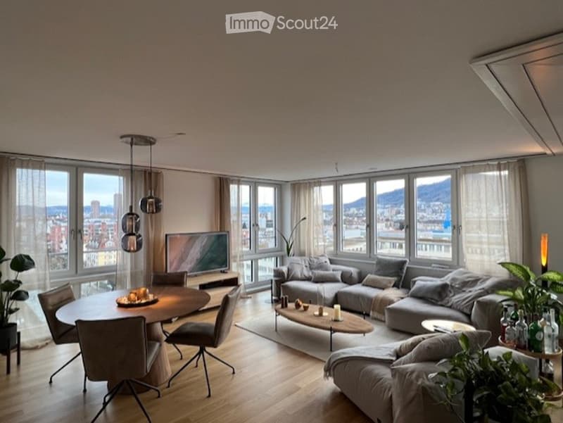 Modern living room with large windows offering a city view, featuring a grey sectional sofa, coffee table, dining table with chairs, and a television. The room has wooden flooring and pendant lights.