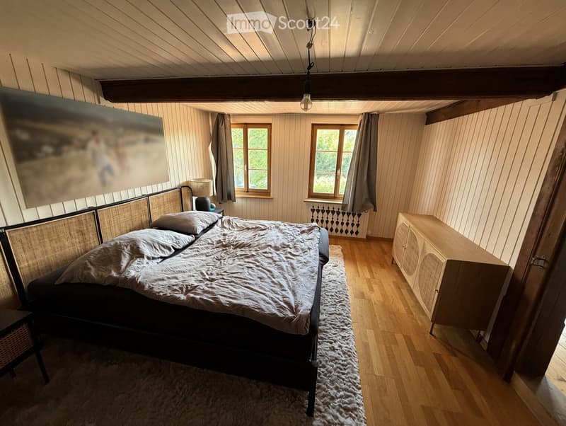 Cozy bedroom with wooden floors, a large bed with a woven headboard, nightstand, and a wooden cabinet. Natural light from two windows with curtains, and a painting on the wall.