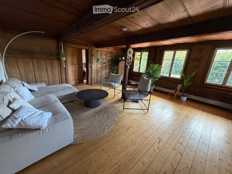 Cozy living room with wooden walls and ceiling, large windows, grey sofa, coffee table, chairs, and plants.
