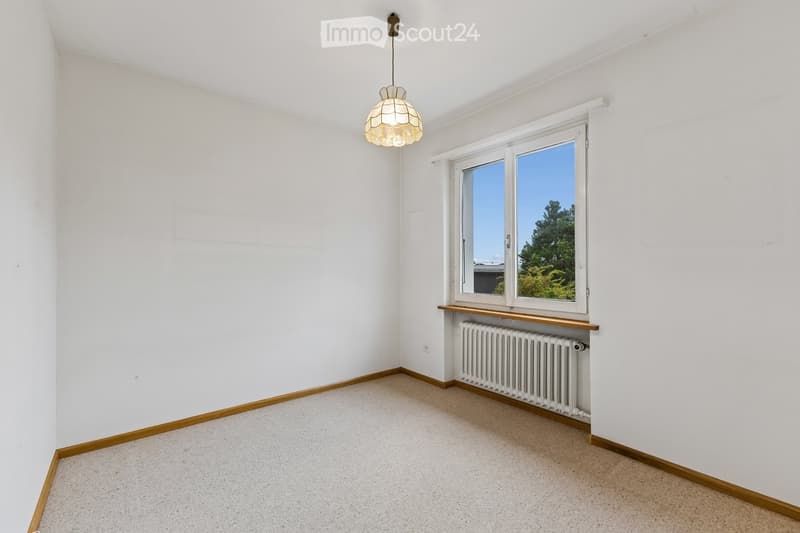 Empty room with white walls, carpeted floor, large window with wooden sill, radiator, and a hanging light fixture.