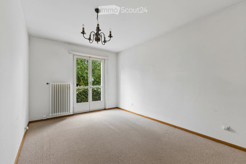 Empty room with white walls, carpeted floor, large white sliding doors leading outside, and a chandelier hanging from the ceiling.