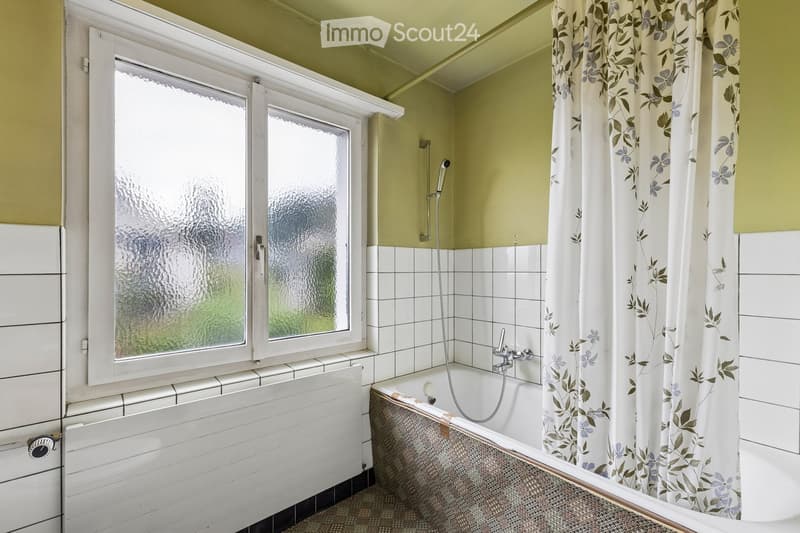 Bathroom with a bathtub, shower curtain, and a window with frosted glass. The walls are tiled, and there is a shower hose attached to the wall.