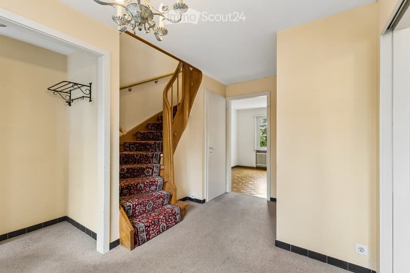 Carpeted hallway with a wooden staircase featuring a red patterned runner and ornate chandelier, leading to an open doorway.