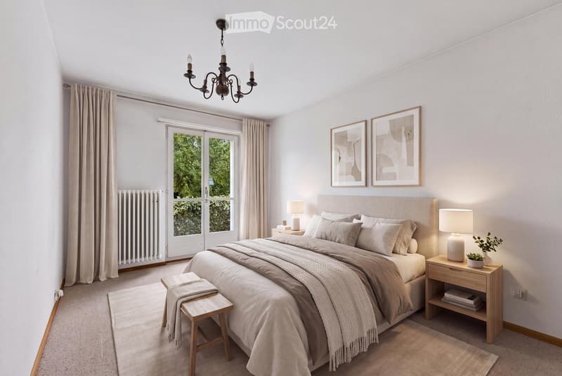 Modern bedroom with a large bed, nightstands, lamps, a wooden stool, framed artwork, and a chandelier. The room features large windows with curtains and a radiator.