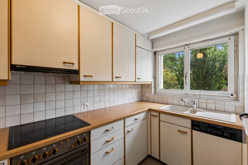 Modern kitchen with light wood cabinets, stainless steel appliances, and a large window overlooking greenery.
