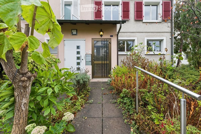 The image shows a garden area leading to the entrance of a two-story building. The pathway is bordered by lush greenery, including shrubs and a tree with large leaves. The building features a white door with a glass panel, a small porch light, and multipl