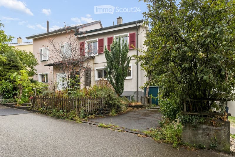 Two-story house with red shutters, surrounded by greenery and a wooden fence.