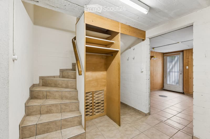 A narrow hallway with white brick walls, tiled floor, wooden built-in shelving unit, staircase leading upwards, and a door at the end.