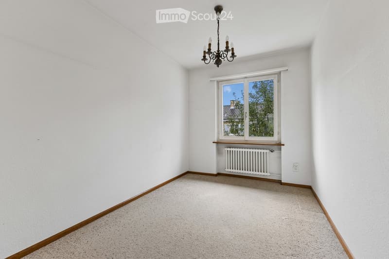 Empty room with white walls, carpeted floor, a window with a view of trees, a radiator, and a chandelier.