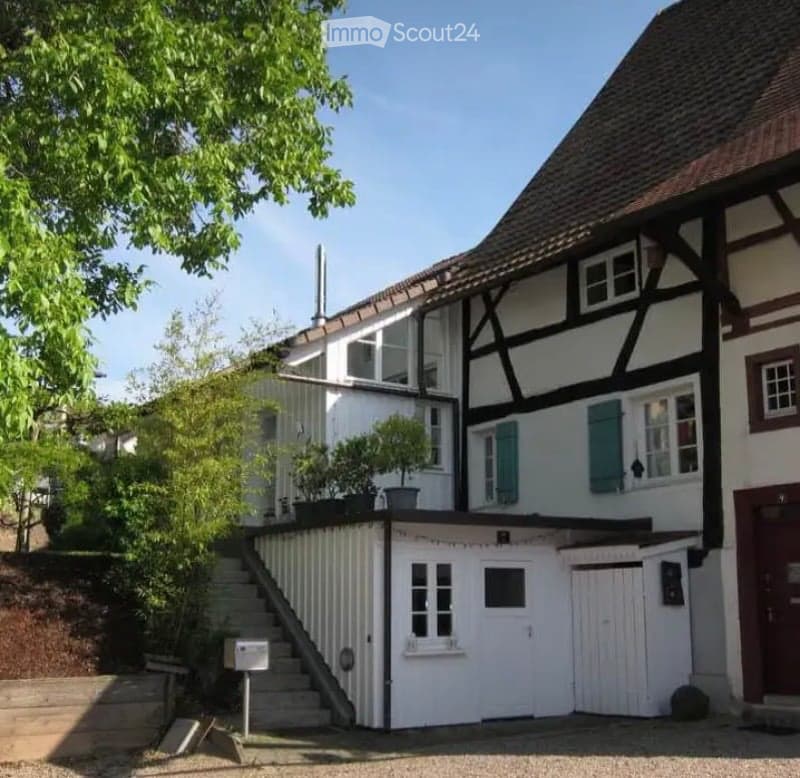 Two-story house with a raised ground floor, white siding, dark wooden beams, and green shutters. A small staircase leads up to the entrance.