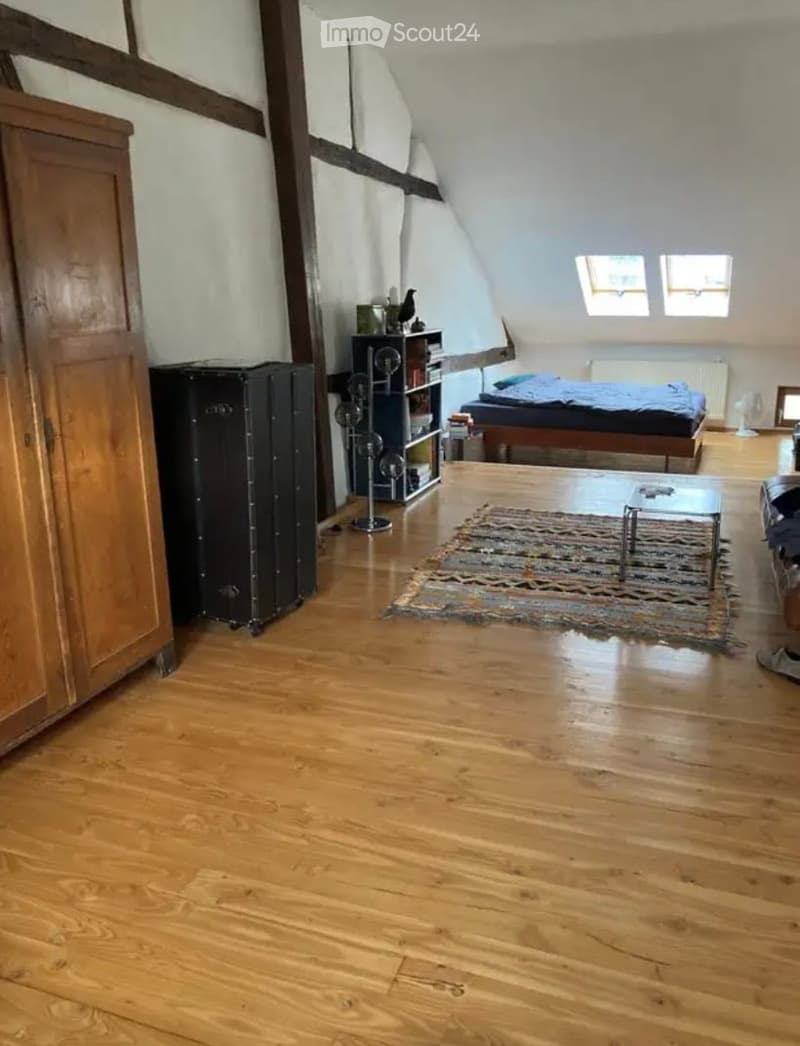 Cozy attic bedroom with wooden floors, slanted ceilings, and skylights. Features a bed, a small table, a rug, a wardrobe, and a bookshelf.