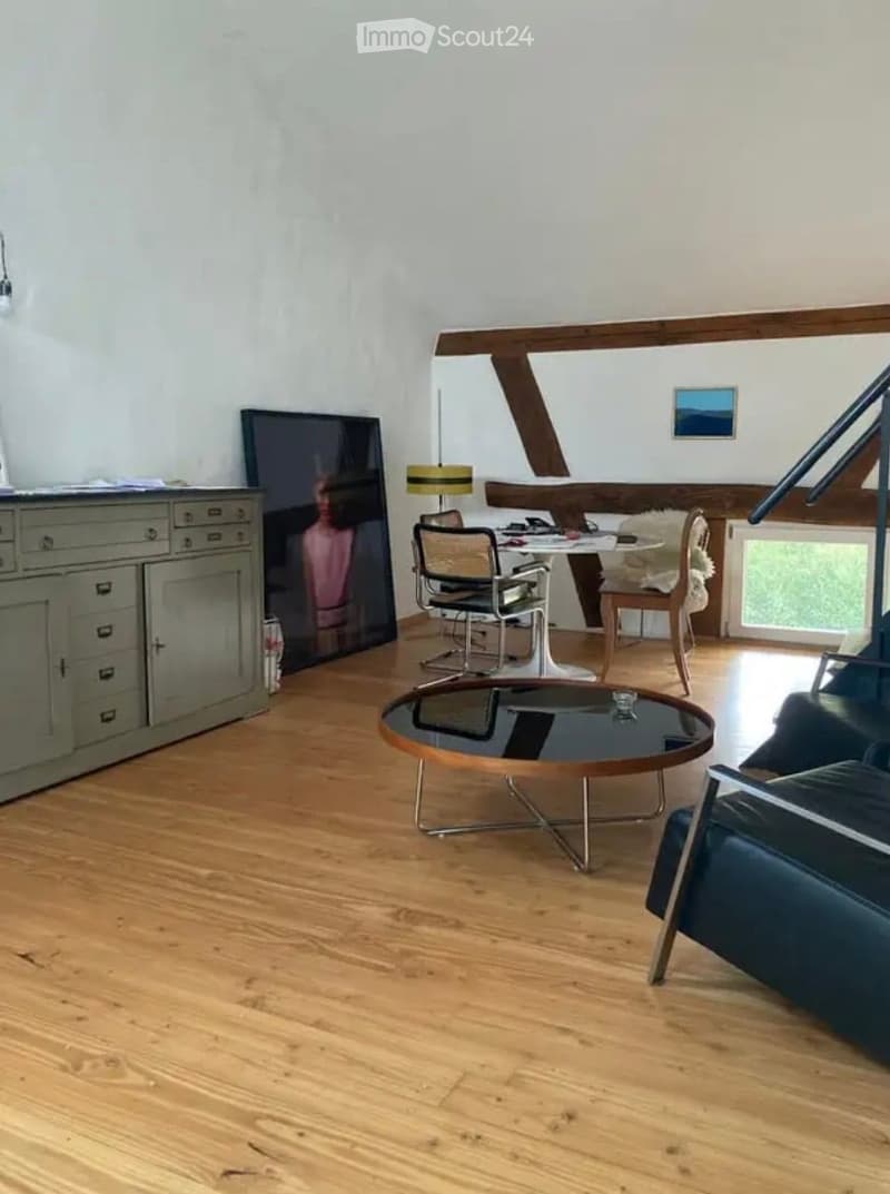 Bright living room with wooden flooring, exposed wooden beams, a sofa, coffee table, dining table with chairs, a cabinet, and a lamp.