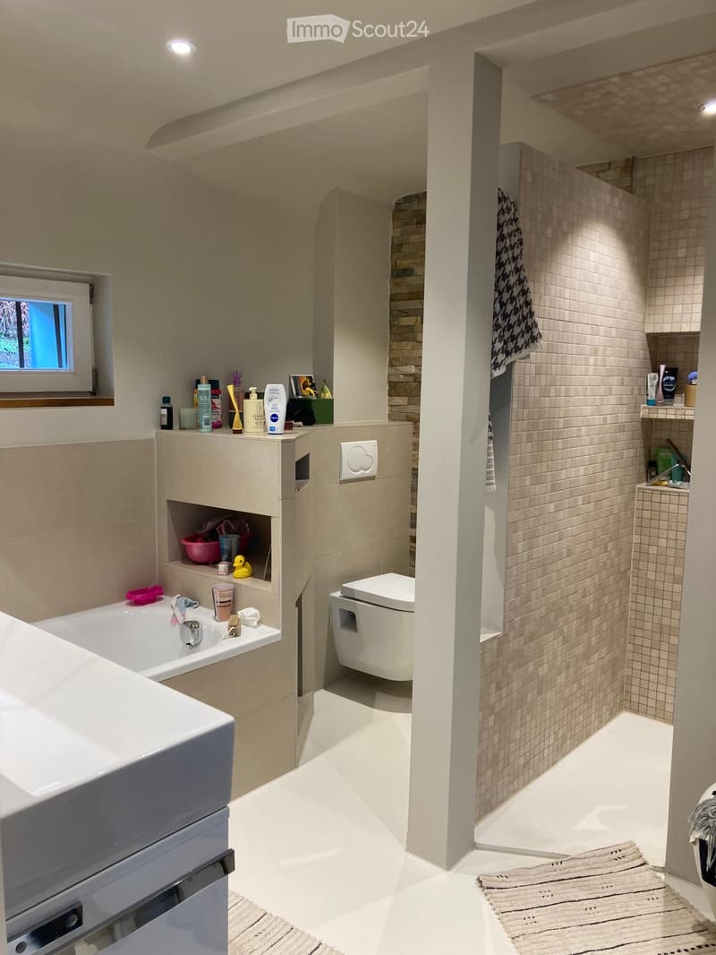 Modern bathroom with a bathtub, toilet, sink, and shower area. Features tiled walls, a window, shelves with toiletries, and a towel hanging on the wall.