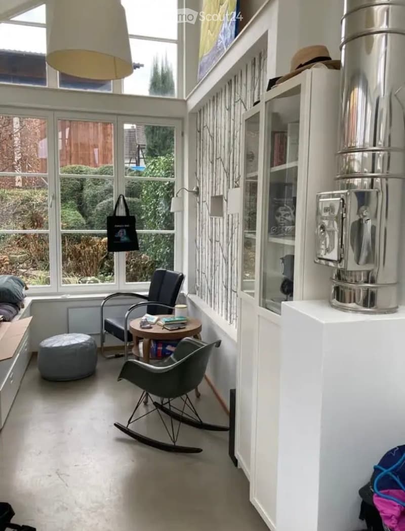Bright, modern living room with large windows, white walls, and a mix of contemporary and classic furniture including a sofa, ottoman, small round table, rocking chair, and a tall cabinet with glass doors.