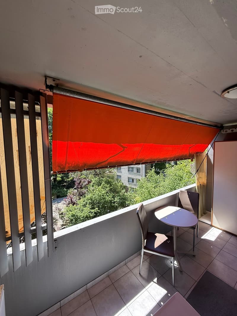 Balcony with tiled floor, table, and chairs, featuring an orange awning and a view of greenery and buildings.