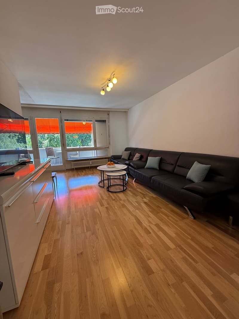 Modern living room with wooden flooring, large windows, a black leather sofa, a small round coffee table, and a television on a stand.