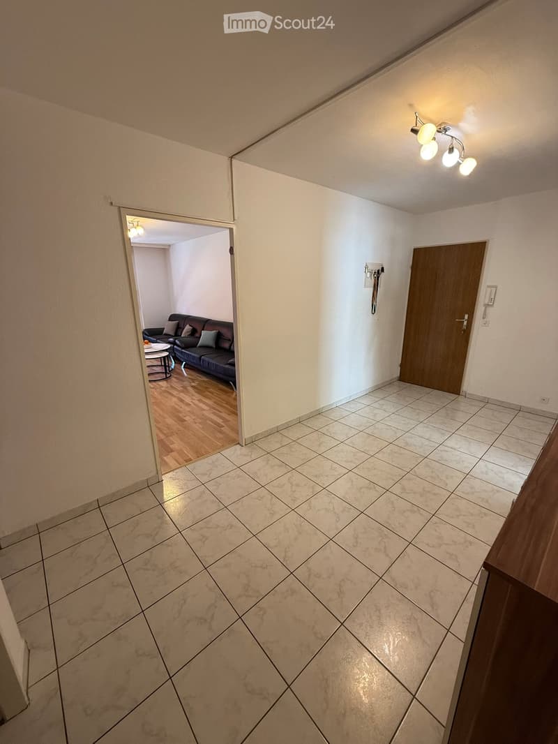 Hallway with tiled floor, white walls, and a wooden door. A chandelier is visible on the ceiling, and an open doorway leads to a living room with a dark-colored sofa and a small round table.