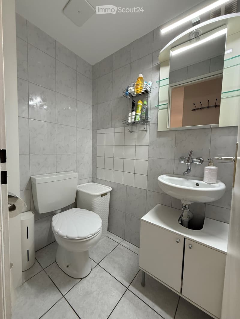 Modern bathroom with white tiles, a toilet, a sink with a mirror, a cabinet, and a shower shelf holding toiletries.