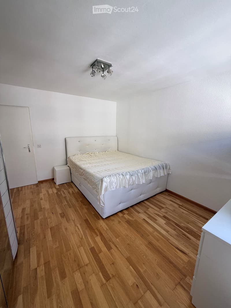 A small bedroom with a single bed, white headboard, and a nightstand. The room features wooden flooring and white walls.