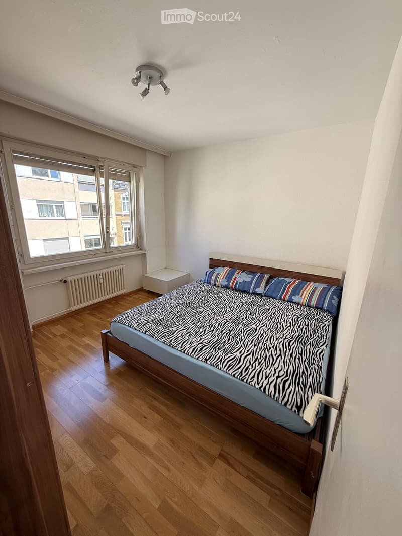 Bedroom with wooden flooring, a single bed with a zebra-patterned duvet and colorful pillows, a nightstand, and a window with blinds.