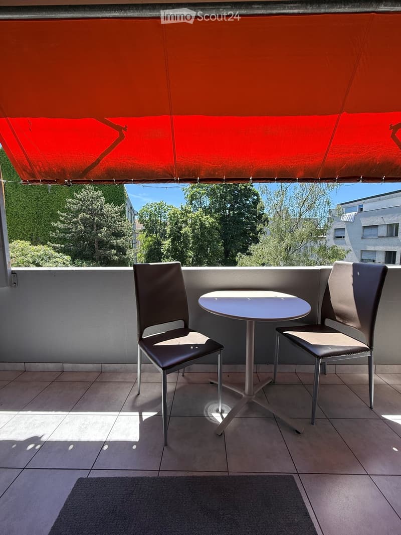 Balcony with a round table and two chairs, covered by a red awning, overlooking a green area with trees and buildings.