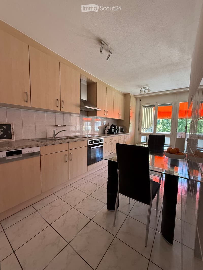 Modern kitchen with light wood cabinets, stainless steel appliances, and a dining area with a glass table and black chairs.