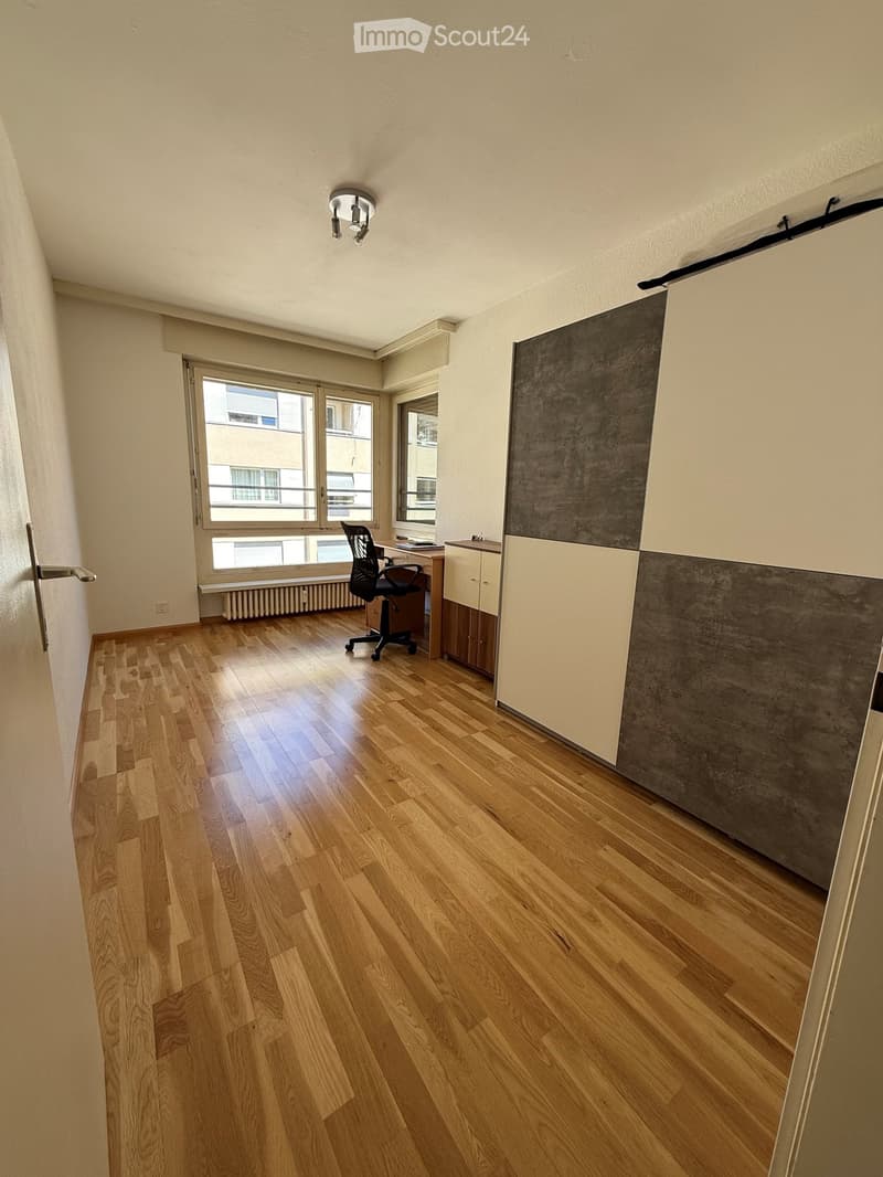 A modern, well-lit room with wooden flooring, a large window, a black office chair, a wooden desk, and a cabinet with a grey and white design.