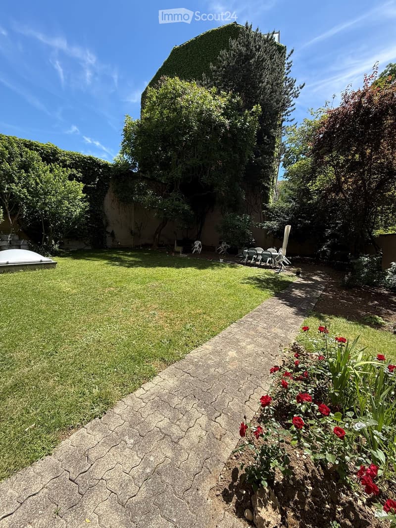Garden with a grassy lawn, stone pathway, small seating area with chairs and table, surrounded by trees and shrubs, with a small dome-shaped structure on the left.