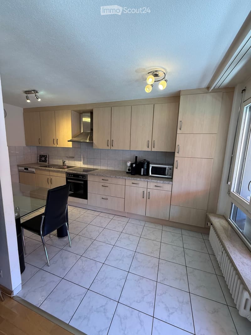Modern kitchen with light wood cabinets, marble tile floor, and stainless steel appliances including an oven, microwave, and dishwasher. A black chair and glass table are positioned near the window.