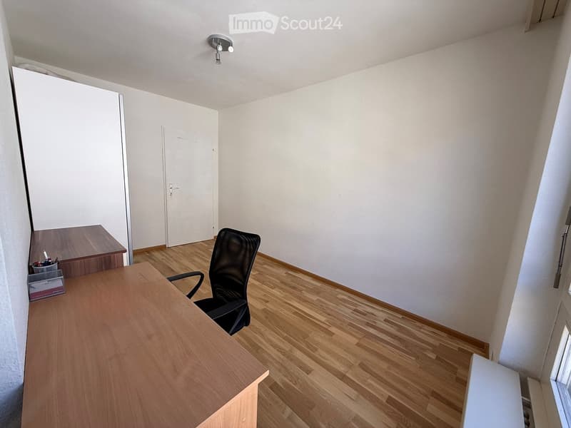 A small, well-lit room with wooden flooring, a large wooden desk, a black office chair, a small cabinet, and a pen holder on the desk.