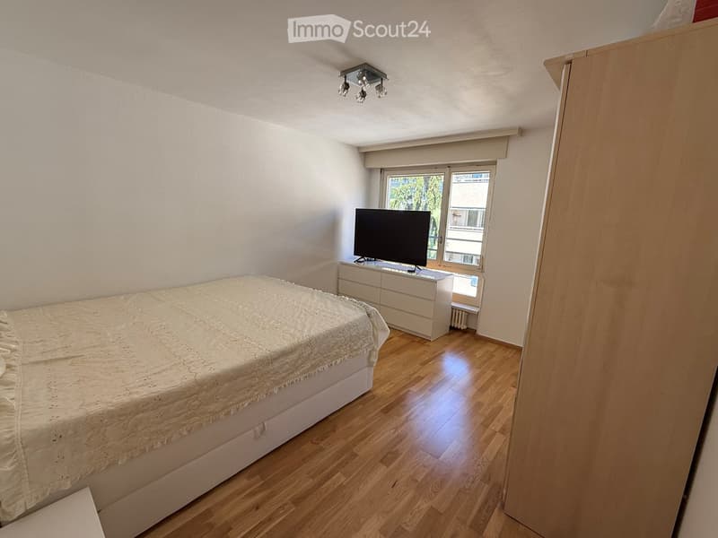 Bedroom with wooden flooring, a neatly made bed, a dresser with a television on top, and a window with a view of greenery.
