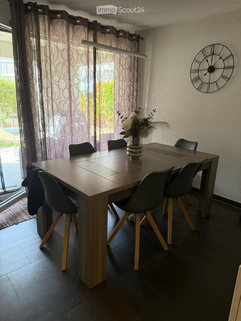 Dining room with a wooden dining table, six black chairs, a vase with flowers, and a large wall clock. The room has dark tiled flooring and large windows with patterned curtains.