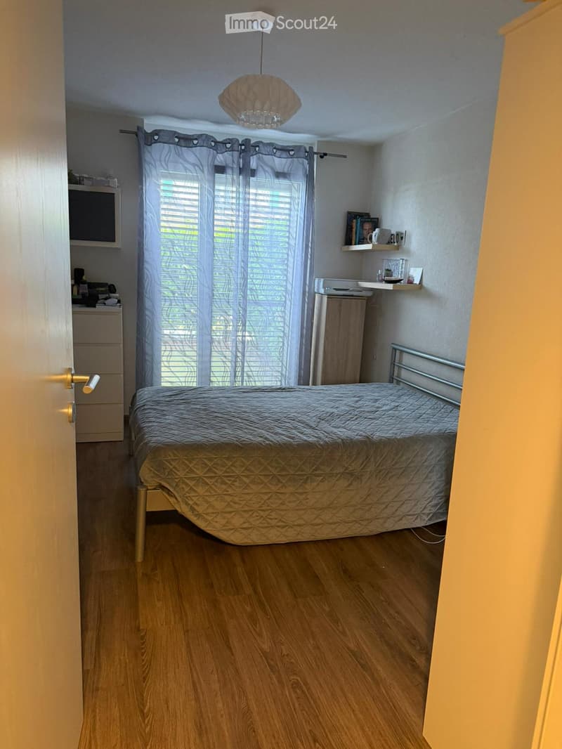 Cozy bedroom with wooden flooring, a single bed with a grey duvet, a nightstand, a shelf with books and a mug, and a window with white curtains.