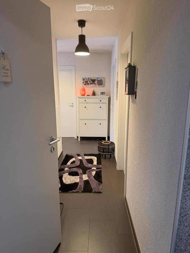 Hallway with tiled floor, white walls, a hanging light fixture, a small white cabinet with drawers, a black and white patterned rug, and a small black stool with a woven design.