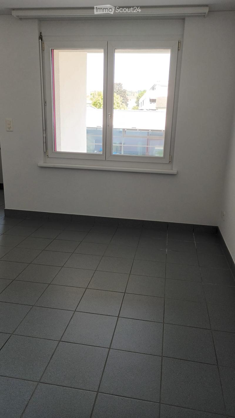 Empty room with grey tiled floor and white walls, featuring a large window with white frame and blinds, and a small electrical outlet on the wall.