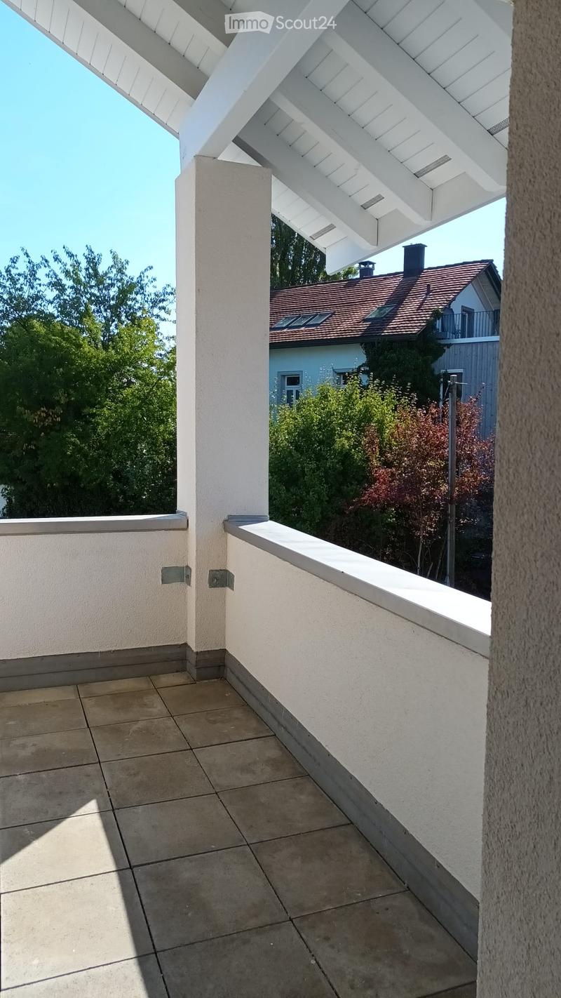 Balcony with tiled floor, white walls, and a white roof structure. The view includes a neighboring house with a red-tiled roof and surrounding greenery.