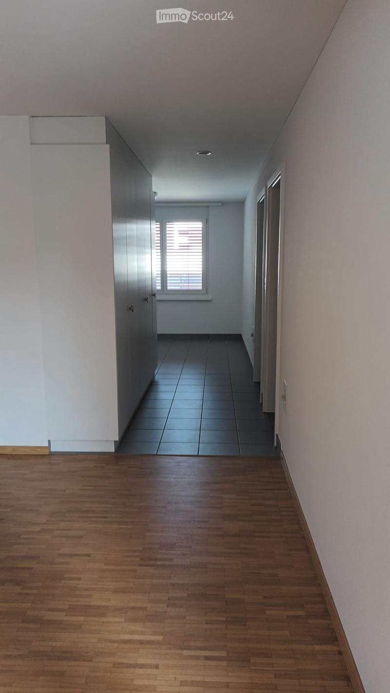 Empty hallway with wooden flooring and tiled section leading to another area, featuring white walls and a window with blinds.