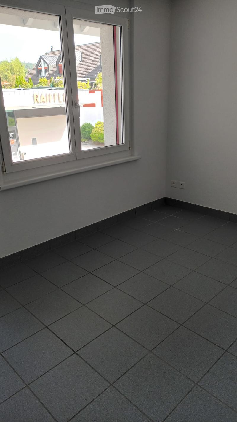 Empty room with grey tiled floor and white walls, featuring a large window with a view of the outside area.