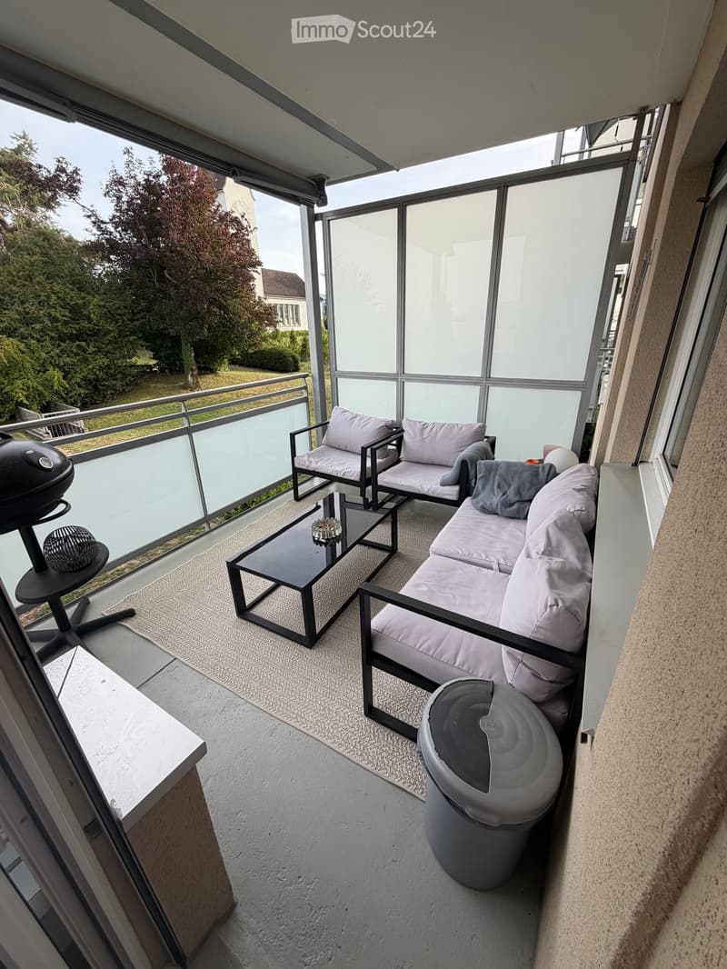 Balcony with modern furniture including a sofa, coffee table, chair, and grill, overlooking a green area with trees and a house.