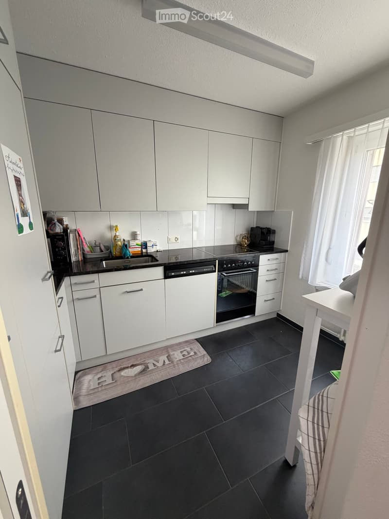 Modern kitchen with white cabinets, black countertops, and black tiled floor. Includes sink, fridge, oven, dishwasher, coffee maker, and a small table.