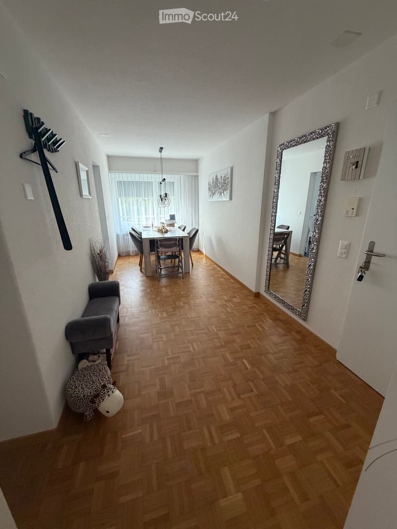 Hallway with wooden flooring, a grey bench, a large mirror, and a small decorative item on the floor. The hallway leads to a dining area with a table and chairs.