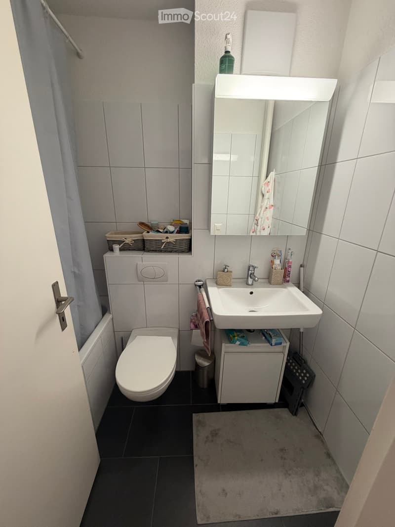 Modern bathroom with white tiles, a toilet, a sink with a mirror, and a wicker basket on the wall holding toiletries.